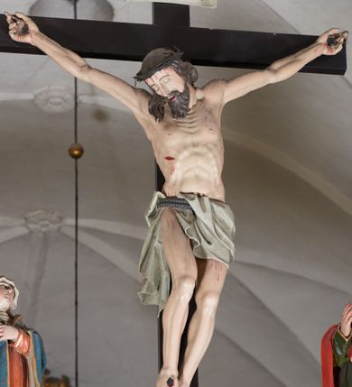 Zdjęcie nr 3: Trójfigurowa grupa Ukrzyżowania, rzeźby pełne. W środku znajduje się krucyfiks z postacią umarłego Chrystusa przybitego trzema gwoździami do krzyża. Chrystus w lekkim zwisie, o szeroko rozłożonych ramionach, bezwładny, głowa opuszczona na prawe ramię. Twarz szczupła, owalna, o prostym nosie, dużych oczach i na wpół otwartych ustach, okolona długim zarostem. Włosy w postaci delikatnych fal opadają na plecy i prawe ramię. Na głowie korona cierniowa. Ciało wychudzone o wyraźnie zarysowanej anatomii. Perizonium w formie tkaniny przewiązanej podwójnym sznurem, odsłaniającej prawe biodro, z dłuższym brzegiem zwisającym po prawej stronie. Draperia gęsto fałdowana, sztywna, o twardych załamaniach materiału. Na szczycie pionowej belki titulus w formie banderoli z napisem „INRI”. Figura Matki Boskiej ustawiona po lewej stronie, zwrócona lekko w stronę krzyża, z głową uniesioną w kierunku Chrystusa. Lewa ręka przyciśnięta do piersi przytrzymuje płaszcz, prawa opuszczona jest wzdłuż ciała. Twarz owalna, niemal bez cech indywidualnych. Ubrana w długą czerwoną suknię, błękitny płaszcz z szarym podbiciem i złoconymi lamówkami, przewiązany diagonalnie przez lewe ramię, opadający na plecy, podtrzymywany na piersi. Na głowie biały welon, opadający na ramiona i plecy. Figura św. Jana Ewangelisty po prawej stronie. Zwrócony delikatnie w stronę krzyża, w kontrapoście, z głową uniesioną w kierunku Chrystusa. Ręce rozpostarte, prawa ugięta i uniesiona w górę, lewa odwiedziona od boku. Twarz młoda, owalna o słabo zindywidualizowanych rysach, włosy w postaci loków sięgają ramion. Święty jest ubrany w ciemnozieloną tunikę przepasaną w talii sznurem oraz czerwony płaszcz ze złoconymi lamówkami, przerzucony diagonalnie przez prawe przedramię i plecy. Matka Boska i św. Jan wykonują podobne gesty; draperie okrywających ich szat są sztywne, ostro załamane. Między figurami Chrystusa i towarzyszącymi mu osobami występuje hierarchia wielkości.