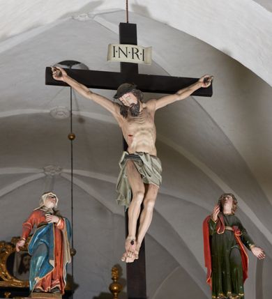 Zdjęcie nr 4: Trójfigurowa grupa Ukrzyżowania, rzeźby pełne. W środku znajduje się krucyfiks z postacią umarłego Chrystusa przybitego trzema gwoździami do krzyża. Chrystus w lekkim zwisie, o szeroko rozłożonych ramionach, bezwładny, głowa opuszczona na prawe ramię. Twarz szczupła, owalna, o prostym nosie, dużych oczach i na wpół otwartych ustach, okolona długim zarostem. Włosy w postaci delikatnych fal opadają na plecy i prawe ramię. Na głowie korona cierniowa. Ciało wychudzone o wyraźnie zarysowanej anatomii. Perizonium w formie tkaniny przewiązanej podwójnym sznurem, odsłaniającej prawe biodro, z dłuższym brzegiem zwisającym po prawej stronie. Draperia gęsto fałdowana, sztywna, o twardych załamaniach materiału. Na szczycie pionowej belki titulus w formie banderoli z napisem „INRI”. Figura Matki Boskiej ustawiona po lewej stronie, zwrócona lekko w stronę krzyża, z głową uniesioną w kierunku Chrystusa. Lewa ręka przyciśnięta do piersi przytrzymuje płaszcz, prawa opuszczona jest wzdłuż ciała. Twarz owalna, niemal bez cech indywidualnych. Ubrana w długą czerwoną suknię, błękitny płaszcz z szarym podbiciem i złoconymi lamówkami, przewiązany diagonalnie przez lewe ramię, opadający na plecy, podtrzymywany na piersi. Na głowie biały welon, opadający na ramiona i plecy. Figura św. Jana Ewangelisty po prawej stronie. Zwrócony delikatnie w stronę krzyża, w kontrapoście, z głową uniesioną w kierunku Chrystusa. Ręce rozpostarte, prawa ugięta i uniesiona w górę, lewa odwiedziona od boku. Twarz młoda, owalna o słabo zindywidualizowanych rysach, włosy w postaci loków sięgają ramion. Święty jest ubrany w ciemnozieloną tunikę przepasaną w talii sznurem oraz czerwony płaszcz ze złoconymi lamówkami, przerzucony diagonalnie przez prawe przedramię i plecy. Matka Boska i św. Jan wykonują podobne gesty; draperie okrywających ich szat są sztywne, ostro załamane. Między figurami Chrystusa i towarzyszącymi mu osobami występuje hierarchia wielkości.