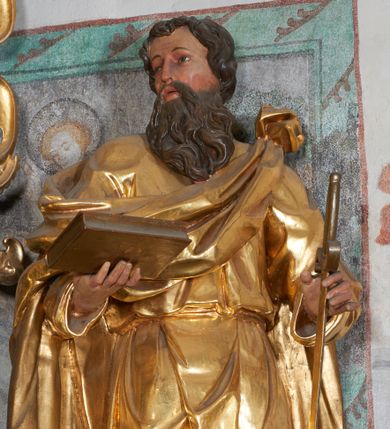 Zdjęcie nr 1: Rzeźba pełna, na niewysokim postumencie, przestawiająca stojącego św. Pawła wyobrażonego w całej postaci. Święty zwrócony frontalnie do widza, z głową skierowaną w prawo, w lekkim kontrapoście; prawa stopa wysunięta do przodu wystaje spod połów szaty. Obie ręce ugięte, wysunięte w przód; w prawej dłoni księga, w lewej miecz skierowany ostrzem w dół. Twarz podłużna, szczupła, z długim, ostrym nosem oraz delikatnie zarysowanymi oczami i ustami, okolona długą brodą, spływającą w postaci pukli na piersi. Włosy krótkie, gęste, kręcone. Święty jest ubrany w tunikę przepasaną w talii sznurem oraz płaszcz przewieszony diagonalnie przez lewe ramię i opadający na plecy. Draperia ekspresyjnie marszczona i fałdowana w partii tuniki, płaszcz w postaci ciężkiej tkaniny obficie drapowanej w grube fałdy opada swobodnie w dół. W partiach ciała polichromia naturalistyczna, szaty złocone, atrybuty polichromowane i delikatnie złocone.