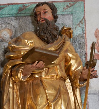 Zdjęcie nr 2: Rzeźba pełna, na niewysokim postumencie, przestawiająca stojącego św. Pawła wyobrażonego w całej postaci. Święty zwrócony frontalnie do widza, z głową skierowaną w prawo, w lekkim kontrapoście; prawa stopa wysunięta do przodu wystaje spod połów szaty. Obie ręce ugięte, wysunięte w przód; w prawej dłoni księga, w lewej miecz skierowany ostrzem w dół. Twarz podłużna, szczupła, z długim, ostrym nosem oraz delikatnie zarysowanymi oczami i ustami, okolona długą brodą, spływającą w postaci pukli na piersi. Włosy krótkie, gęste, kręcone. Święty jest ubrany w tunikę przepasaną w talii sznurem oraz płaszcz przewieszony diagonalnie przez lewe ramię i opadający na plecy. Draperia ekspresyjnie marszczona i fałdowana w partii tuniki, płaszcz w postaci ciężkiej tkaniny obficie drapowanej w grube fałdy opada swobodnie w dół. W partiach ciała polichromia naturalistyczna, szaty złocone, atrybuty polichromowane i delikatnie złocone.