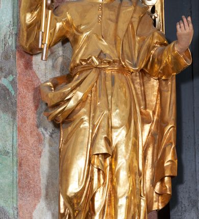 Zdjęcie nr 2: Rzeźba pełna, na niewysokim postumencie, przestawiająca św. Piotra. Święty zwrócony frontalnie do widza, z głową skręconą nieznacznie w lewo, ukazany w lekkim kontrapoście; lewa stopa wysunięta do przodu wystaje spod szaty. Obie ręce zgięte, uniesione, w prawej dłoni dwa klucze, lewa zwrócona ku górze. Twarz pociągła, szczupła, z dużym, ostrym nosem oraz delikatnie zarysowanymi ustami i oczami. Okolona stosunkowo krótką, falowaną brodą; włosy krótkie w formie pukli. Święty jest ubrany w długą tunikę, zapinaną pod szyją na guziki, przepasaną sznurem w talii oraz płaszcz przewieszony przez lewe ramię, opadający przez plecy, zawinięty wokół prawego biodra i zatknięty z przodu za pasek. Draperia ukształtowana miękko, o delikatnych fałdowaniach tuniki oraz grubych załamaniach ciężkiej tkaniny płaszcza. Polichromia w partiach ciała naturalistyczna, szaty i atrybuty złocone.