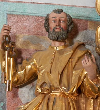 Zdjęcie nr 3: Rzeźba pełna, na niewysokim postumencie, przestawiająca św. Piotra. Święty zwrócony frontalnie do widza, z głową skręconą nieznacznie w lewo, ukazany w lekkim kontrapoście; lewa stopa wysunięta do przodu wystaje spod szaty. Obie ręce zgięte, uniesione, w prawej dłoni dwa klucze, lewa zwrócona ku górze. Twarz pociągła, szczupła, z dużym, ostrym nosem oraz delikatnie zarysowanymi ustami i oczami. Okolona stosunkowo krótką, falowaną brodą; włosy krótkie w formie pukli. Święty jest ubrany w długą tunikę, zapinaną pod szyją na guziki, przepasaną sznurem w talii oraz płaszcz przewieszony przez lewe ramię, opadający przez plecy, zawinięty wokół prawego biodra i zatknięty z przodu za pasek. Draperia ukształtowana miękko, o delikatnych fałdowaniach tuniki oraz grubych załamaniach ciężkiej tkaniny płaszcza. Polichromia w partiach ciała naturalistyczna, szaty i atrybuty złocone.