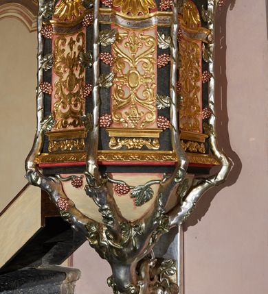 Zdjęcie nr 3: Ambona przyścienna, ustawiona na rzeźbionym konarze, rozwarstwiającym się na pnącza ujmujące kosz. Kosz na planie ośmioboku na cokoliku, u spodu podwieszona półkula. Ścianki wydzielone rzeźbionymi pnączami z winnym gronem. W polach płytkie nisze, wsparte na cokolikach, wypełnione okuciami przechodzącymi w wić roślinną, u góry zamknięte półkoliście z konchami. Ścianki w dwóch trzecich wysokości włącznie z wnękami obiega profilowany gzyms. Parapet kosza w formie gzymsu dekorowany głębokim kimationem. Zaplecek o kształcie stojącego prostokąta wypełnionego kanelurami, zamkniętego fryzem z metopami zdobionymi kulami; ujęty strzępionymi liśćmi palmowymi. Baldachim na planie analogicznym względem kosza, płaski, zwieńczony palmetami; na podniebiu rzeźbiona gołębica Ducha Świętego w medalionie utworzonym z liśćmi wawrzynu, w glorii promienistej. Struktura polichromowana na kolor czarny, podwieszenie kosza na kolor biały; ornamentyka, profilowania i detale złocone, srebrzone oraz polichromowane na czerwono. Ornamentyka na koszu dodatkowo pokryta laserunkiem. 