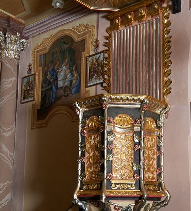 Zdjęcie nr 1: Ambona przyścienna, ustawiona na rzeźbionym konarze, rozwarstwiającym się na pnącza ujmujące kosz. Kosz na planie ośmioboku na cokoliku, u spodu podwieszona półkula. Ścianki wydzielone rzeźbionymi pnączami z winnym gronem. W polach płytkie nisze, wsparte na cokolikach, wypełnione okuciami przechodzącymi w wić roślinną, u góry zamknięte półkoliście z konchami. Ścianki w dwóch trzecich wysokości włącznie z wnękami obiega profilowany gzyms. Parapet kosza w formie gzymsu dekorowany głębokim kimationem. Zaplecek o kształcie stojącego prostokąta wypełnionego kanelurami, zamkniętego fryzem z metopami zdobionymi kulami; ujęty strzępionymi liśćmi palmowymi. Baldachim na planie analogicznym względem kosza, płaski, zwieńczony palmetami; na podniebiu rzeźbiona gołębica Ducha Świętego w medalionie utworzonym z liśćmi wawrzynu, w glorii promienistej. Struktura polichromowana na kolor czarny, podwieszenie kosza na kolor biały; ornamentyka, profilowania i detale złocone, srebrzone oraz polichromowane na czerwono. Ornamentyka na koszu dodatkowo pokryta laserunkiem. 