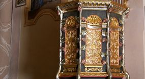 Zdjęcie nr 1: Ambona przyścienna, ustawiona na rzeźbionym konarze, rozwarstwiającym się na pnącza ujmujące kosz. Kosz na planie ośmioboku na cokoliku, u spodu podwieszona półkula. Ścianki wydzielone rzeźbionymi pnączami z winnym gronem. W polach płytkie nisze, wsparte na cokolikach, wypełnione okuciami przechodzącymi w wić roślinną, u góry zamknięte półkoliście z konchami. Ścianki w dwóch trzecich wysokości włącznie z wnękami obiega profilowany gzyms. Parapet kosza w formie gzymsu dekorowany głębokim kimationem. Zaplecek o kształcie stojącego prostokąta wypełnionego kanelurami, zamkniętego fryzem z metopami zdobionymi kulami; ujęty strzępionymi liśćmi palmowymi. Baldachim na planie analogicznym względem kosza, płaski, zwieńczony palmetami; na podniebiu rzeźbiona gołębica Ducha Świętego w medalionie utworzonym z liśćmi wawrzynu, w glorii promienistej. Struktura polichromowana na kolor czarny, podwieszenie kosza na kolor biały; ornamentyka, profilowania i detale złocone, srebrzone oraz polichromowane na czerwono. Ornamentyka na koszu dodatkowo pokryta laserunkiem. 