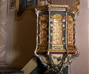 Zdjęcie nr 1: Ambona przyścienna, ustawiona na rzeźbionym konarze, rozwarstwiającym się na pnącza ujmujące kosz. Kosz na planie ośmioboku na cokoliku, u spodu podwieszona półkula. Ścianki wydzielone rzeźbionymi pnączami z winnym gronem. W polach płytkie nisze, wsparte na cokolikach, wypełnione okuciami przechodzącymi w wić roślinną, u góry zamknięte półkoliście z konchami. Ścianki w dwóch trzecich wysokości włącznie z wnękami obiega profilowany gzyms. Parapet kosza w formie gzymsu dekorowany głębokim kimationem. Zaplecek o kształcie stojącego prostokąta wypełnionego kanelurami, zamkniętego fryzem z metopami zdobionymi kulami; ujęty strzępionymi liśćmi palmowymi. Baldachim na planie analogicznym względem kosza, płaski, zwieńczony palmetami; na podniebiu rzeźbiona gołębica Ducha Świętego w medalionie utworzonym z liśćmi wawrzynu, w glorii promienistej. Struktura polichromowana na kolor czarny, podwieszenie kosza na kolor biały; ornamentyka, profilowania i detale złocone, srebrzone oraz polichromowane na czerwono. Ornamentyka na koszu dodatkowo pokryta laserunkiem. 