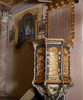 Zdjęcie nr 1: Ambona przyścienna, ustawiona na rzeźbionym konarze, rozwarstwiającym się na pnącza ujmujące kosz. Kosz na planie ośmioboku na cokoliku, u spodu podwieszona półkula. Ścianki wydzielone rzeźbionymi pnączami z winnym gronem. W polach płytkie nisze, wsparte na cokolikach, wypełnione okuciami przechodzącymi w wić roślinną, u góry zamknięte półkoliście z konchami. Ścianki w dwóch trzecich wysokości włącznie z wnękami obiega profilowany gzyms. Parapet kosza w formie gzymsu dekorowany głębokim kimationem. Zaplecek o kształcie stojącego prostokąta wypełnionego kanelurami, zamkniętego fryzem z metopami zdobionymi kulami; ujęty strzępionymi liśćmi palmowymi. Baldachim na planie analogicznym względem kosza, płaski, zwieńczony palmetami; na podniebiu rzeźbiona gołębica Ducha Świętego w medalionie utworzonym z liśćmi wawrzynu, w glorii promienistej. Struktura polichromowana na kolor czarny, podwieszenie kosza na kolor biały; ornamentyka, profilowania i detale złocone, srebrzone oraz polichromowane na czerwono. Ornamentyka na koszu dodatkowo pokryta laserunkiem. 