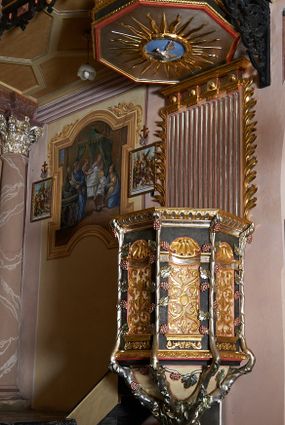 Zdjęcie nr 1: Ambona przyścienna, ustawiona na rzeźbionym konarze, rozwarstwiającym się na pnącza ujmujące kosz. Kosz na planie ośmioboku na cokoliku, u spodu podwieszona półkula. Ścianki wydzielone rzeźbionymi pnączami z winnym gronem. W polach płytkie nisze, wsparte na cokolikach, wypełnione okuciami przechodzącymi w wić roślinną, u góry zamknięte półkoliście z konchami. Ścianki w dwóch trzecich wysokości włącznie z wnękami obiega profilowany gzyms. Parapet kosza w formie gzymsu dekorowany głębokim kimationem. Zaplecek o kształcie stojącego prostokąta wypełnionego kanelurami, zamkniętego fryzem z metopami zdobionymi kulami; ujęty strzępionymi liśćmi palmowymi. Baldachim na planie analogicznym względem kosza, płaski, zwieńczony palmetami; na podniebiu rzeźbiona gołębica Ducha Świętego w medalionie utworzonym z liśćmi wawrzynu, w glorii promienistej. Struktura polichromowana na kolor czarny, podwieszenie kosza na kolor biały; ornamentyka, profilowania i detale złocone, srebrzone oraz polichromowane na czerwono. Ornamentyka na koszu dodatkowo pokryta laserunkiem. 