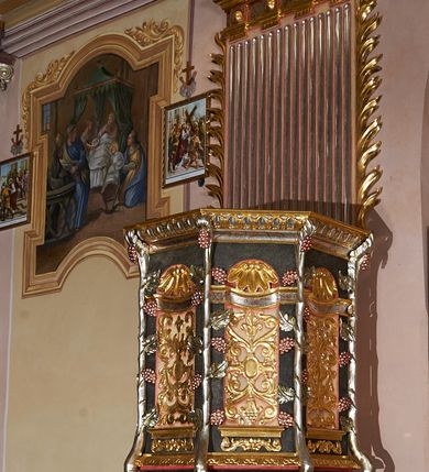 Zdjęcie nr 2: Ambona przyścienna, ustawiona na rzeźbionym konarze, rozwarstwiającym się na pnącza ujmujące kosz. Kosz na planie ośmioboku na cokoliku, u spodu podwieszona półkula. Ścianki wydzielone rzeźbionymi pnączami z winnym gronem. W polach płytkie nisze, wsparte na cokolikach, wypełnione okuciami przechodzącymi w wić roślinną, u góry zamknięte półkoliście z konchami. Ścianki w dwóch trzecich wysokości włącznie z wnękami obiega profilowany gzyms. Parapet kosza w formie gzymsu dekorowany głębokim kimationem. Zaplecek o kształcie stojącego prostokąta wypełnionego kanelurami, zamkniętego fryzem z metopami zdobionymi kulami; ujęty strzępionymi liśćmi palmowymi. Baldachim na planie analogicznym względem kosza, płaski, zwieńczony palmetami; na podniebiu rzeźbiona gołębica Ducha Świętego w medalionie utworzonym z liśćmi wawrzynu, w glorii promienistej. Struktura polichromowana na kolor czarny, podwieszenie kosza na kolor biały; ornamentyka, profilowania i detale złocone, srebrzone oraz polichromowane na czerwono. Ornamentyka na koszu dodatkowo pokryta laserunkiem. 