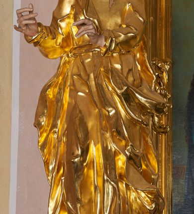 Zdjęcie nr 3: Rzeźba drążona, ustawiona na prostopadłościennym cokole. Figura ukazana frontalnie, w silnym kontrapoście z prawą nogą ugiętą w kolanie, z głową skierowaną w lewo, z dwoma rękami wyciągniętymi w prawą stronę. Twarz szczupła, pociągła, wyraźnie zarysowane linie łuków brwiowych, nosa i kości policzkowych; oczy niewielkie, głęboko osadzone, nos wąski, prosty, usta pełne, rozchylone; okolona krótką, kręconą brodą. Włosy krótkie, głęboko rzeźbione w pojedyncze pukle, opadające na plecy. Święty ubrany jest w suknię rozchyloną na piersi, z kołnierzykiem; przepasaną szarfą w talii; rękawy długie z podwiniętym brzegiem; tkanina ściśle przylegająca do ciała, marszczona, silnie drapowana. Płaszcz przerzucony przez lewe ramię, otaczający postać i zawinięty na prawym boku, z fragmentem upiętym u pasa; tkanina silnie drapowana, o wywiniętym brzegu i rozwianych fragmentach. Polichromia naturalistyczna w odsłoniętych partiach ciała, szata złocona.