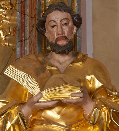Zdjęcie nr 9: Rzeźba drążona, ustawiona na cokole. Figura ukazana frontalnie, w kontrapoście, obiema rękami podtrzymuje przed piersią otwartą księgę. Twarz pociągła, o wydłużonej szczęce; oczy duże, spoglądające w dół, nos prosty i duży, usta pełne; okolona krótką, kręconą brodą. Włosy krótkie, falowane. Święty ubrany jest w długą suknię z kołnierzykiem, z długimi rękawami o podwiniętym brzegu, przepasaną w talii szarfą; tkanina ściśle przylega do ciała, w partii torsu marszczona, delikatnie drapowana w zygzakowate fałdki w dolnej części. Płaszcz podtrzymywany na przedramionach, z obfitą partią materiału zawijającą się na lewej nodze; tkanina delikatnie rozwiana, miejscami zawinięta, silnie drapowana. Polichromia naturalistyczna w odsłoniętych partiach ciała, szaty i atrybuty złocone.