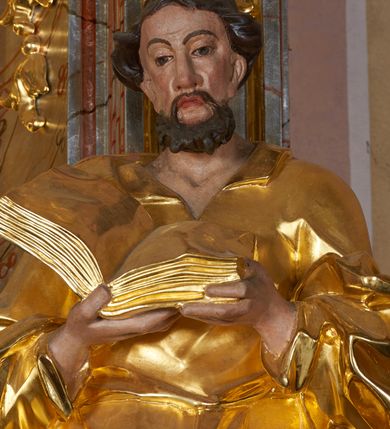 Zdjęcie nr 11: Rzeźba drążona, ustawiona na cokole. Figura ukazana frontalnie, w kontrapoście, obiema rękami podtrzymuje przed piersią otwartą księgę. Twarz pociągła, o wydłużonej szczęce; oczy duże, spoglądające w dół, nos prosty i duży, usta pełne; okolona krótką, kręconą brodą. Włosy krótkie, falowane. Święty ubrany jest w długą suknię z kołnierzykiem, z długimi rękawami o podwiniętym brzegu, przepasaną w talii szarfą; tkanina ściśle przylega do ciała, w partii torsu marszczona, delikatnie drapowana w zygzakowate fałdki w dolnej części. Płaszcz podtrzymywany na przedramionach, z obfitą partią materiału zawijającą się na lewej nodze; tkanina delikatnie rozwiana, miejscami zawinięta, silnie drapowana. Polichromia naturalistyczna w odsłoniętych partiach ciała, szaty i atrybuty złocone.