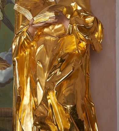 Zdjęcie nr 4: Rzeźba drążona, ustawiona na cokole. Figura ukazana frontalnie, w kontrapoście, obiema rękami podtrzymuje przed piersią otwartą księgę. Twarz pociągła, o wydłużonej szczęce; oczy duże, spoglądające w dół, nos prosty i duży, usta pełne; okolona krótką, kręconą brodą. Włosy krótkie, falowane. Święty ubrany jest w długą suknię z kołnierzykiem, z długimi rękawami o podwiniętym brzegu, przepasaną w talii szarfą; tkanina ściśle przylega do ciała, w partii torsu marszczona, delikatnie drapowana w zygzakowate fałdki w dolnej części. Płaszcz podtrzymywany na przedramionach, z obfitą partią materiału zawijającą się na lewej nodze; tkanina delikatnie rozwiana, miejscami zawinięta, silnie drapowana. Polichromia naturalistyczna w odsłoniętych partiach ciała, szaty i atrybuty złocone.