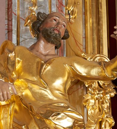 Zdjęcie nr 3: Rzeźba drążona, ustawiona na cokole. Figura ukazana frontalnie, w kontrapoście z prawą nogą ugiętą w kolanie, z uniesioną, skierowaną w lewo głową; lewa ręka wyciągnięta w bok, a prawa podtrzymuje na boku zamkniętą księgę. Twarz szczupła, pociągła, z wysokim czołem, o podkreślonych liniach łuków brwiowych, nosa i kości policzkowych, oczy głęboko osadzone, zwrócone ku górze, usta pełne, rozchylone; okolona krótką i kręconą brodą. Włosy rzeźbione w pojedyncze pukle, usytuowane po bokach i z tyłu głowy. Święty ubrany jest w suknię o wąskich rękawach, z wywiniętym brzegiem, ściśle przylegającą do ciała; tkanina drapowana jest w pojedyncze fałdki, u spodu w grube, głęboko rzeźbione. Na prawe ramię święty ma narzucony płaszcz, opasujący postać, zawieszony na lewym biodrze; tkanina jest ekspresyjnie drapowana, poruszona. Polichromia naturalistyczna w odsłoniętych partiach ciała, szaty i atrybuty złocone.