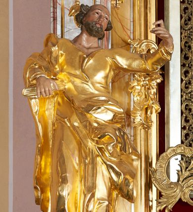 Zdjęcie nr 1: Rzeźba drążona, ustawiona na cokole. Figura ukazana frontalnie, w kontrapoście z prawą nogą ugiętą w kolanie, z uniesioną, skierowaną w lewo głową; lewa ręka wyciągnięta w bok, a prawa podtrzymuje na boku zamkniętą księgę. Twarz szczupła, pociągła, z wysokim czołem, o podkreślonych liniach łuków brwiowych, nosa i kości policzkowych, oczy głęboko osadzone, zwrócone ku górze, usta pełne, rozchylone; okolona krótką i kręconą brodą. Włosy rzeźbione w pojedyncze pukle, usytuowane po bokach i z tyłu głowy. Święty ubrany jest w suknię o wąskich rękawach, z wywiniętym brzegiem, ściśle przylegającą do ciała; tkanina drapowana jest w pojedyncze fałdki, u spodu w grube, głęboko rzeźbione. Na prawe ramię święty ma narzucony płaszcz, opasujący postać, zawieszony na lewym biodrze; tkanina jest ekspresyjnie drapowana, poruszona. Polichromia naturalistyczna w odsłoniętych partiach ciała, szaty i atrybuty złocone.