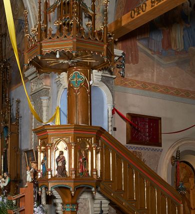 Zdjęcie nr 1: Ambona przyścienna, składająca się z kosza, zaplecka i baldachimu, z dostępem od prawej strony w postaci jednobiegowych schodów z balustradą. Kosz na planie ośmioboku, ścianki dekorowane płycinami wydzielonymi przez półkolumny o kapitelach z dekoracją kwiatową na wielobocznych postumentach podtrzymujących belkowanie, którego wysuniety gzyms stanowi parapet. W polach blendy z płaskorzeźbionymi przedstawieniami czterech ewangelistów. Każdy z nich został ukazany całopostaciowo, w długiej szacie, z księgą lub zwojem w ręce i w towarzystwie symbolu apokaliptycznego. W przyłuczach blend ostrokrzew. Kosz z podwieszeniem z guzów, oparty na ośmiobocznym filarze zwieńczonym pierścieniem z dekoracją w formie rozet, ustawiony na profilowanym cokole. Balustrada dekorowana ostrołukowymi blendami. Zaplecek w kształcie wydłużonego stojącego prostokąta, półokrągły. Dekorowany płyciną z parą blend i czworoliściem z krzyżem równoramiennym. Baldachim ośmioboczny, zwieńczony ażurową wieżyczką z wimpergami, wspartą czterema przyporami, dekorowaną pinaklami, żabkami i astwerkiem; zwieńczoną pinaklem z krzyżem. Na podniebiu płaskorzeźbione przedstawienie gołębicy Ducha Świętego w glorii promienistej. Całość struktury w odcieniu ciemnego drewna, profile i detale niebieskie i czerwone, reszta profilów, wnęki blend z płaskorzeźbami i detale złocone. 