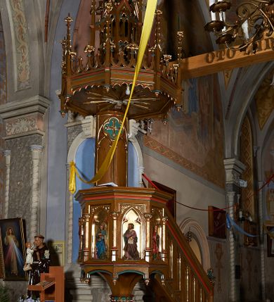 Zdjęcie nr 4: Ambona przyścienna, składająca się z kosza, zaplecka i baldachimu, z dostępem od prawej strony w postaci jednobiegowych schodów z balustradą. Kosz na planie ośmioboku, ścianki dekorowane płycinami wydzielonymi przez półkolumny o kapitelach z dekoracją kwiatową na wielobocznych postumentach podtrzymujących belkowanie, którego wysuniety gzyms stanowi parapet. W polach blendy z płaskorzeźbionymi przedstawieniami czterech ewangelistów. Każdy z nich został ukazany całopostaciowo, w długiej szacie, z księgą lub zwojem w ręce i w towarzystwie symbolu apokaliptycznego. W przyłuczach blend ostrokrzew. Kosz z podwieszeniem z guzów, oparty na ośmiobocznym filarze zwieńczonym pierścieniem z dekoracją w formie rozet, ustawiony na profilowanym cokole. Balustrada dekorowana ostrołukowymi blendami. Zaplecek w kształcie wydłużonego stojącego prostokąta, półokrągły. Dekorowany płyciną z parą blend i czworoliściem z krzyżem równoramiennym. Baldachim ośmioboczny, zwieńczony ażurową wieżyczką z wimpergami, wspartą czterema przyporami, dekorowaną pinaklami, żabkami i astwerkiem; zwieńczoną pinaklem z krzyżem. Na podniebiu płaskorzeźbione przedstawienie gołębicy Ducha Świętego w glorii promienistej. Całość struktury w odcieniu ciemnego drewna, profile i detale niebieskie i czerwone, reszta profilów, wnęki blend z płaskorzeźbami i detale złocone. 