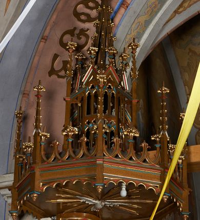 Zdjęcie nr 6: Ambona przyścienna, składająca się z kosza, zaplecka i baldachimu, z dostępem od prawej strony w postaci jednobiegowych schodów z balustradą. Kosz na planie ośmioboku, ścianki dekorowane płycinami wydzielonymi przez półkolumny o kapitelach z dekoracją kwiatową na wielobocznych postumentach podtrzymujących belkowanie, którego wysuniety gzyms stanowi parapet. W polach blendy z płaskorzeźbionymi przedstawieniami czterech ewangelistów. Każdy z nich został ukazany całopostaciowo, w długiej szacie, z księgą lub zwojem w ręce i w towarzystwie symbolu apokaliptycznego. W przyłuczach blend ostrokrzew. Kosz z podwieszeniem z guzów, oparty na ośmiobocznym filarze zwieńczonym pierścieniem z dekoracją w formie rozet, ustawiony na profilowanym cokole. Balustrada dekorowana ostrołukowymi blendami. Zaplecek w kształcie wydłużonego stojącego prostokąta, półokrągły. Dekorowany płyciną z parą blend i czworoliściem z krzyżem równoramiennym. Baldachim ośmioboczny, zwieńczony ażurową wieżyczką z wimpergami, wspartą czterema przyporami, dekorowaną pinaklami, żabkami i astwerkiem; zwieńczoną pinaklem z krzyżem. Na podniebiu płaskorzeźbione przedstawienie gołębicy Ducha Świętego w glorii promienistej. Całość struktury w odcieniu ciemnego drewna, profile i detale niebieskie i czerwone, reszta profilów, wnęki blend z płaskorzeźbami i detale złocone. 