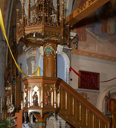 Zdjęcie nr 2: Ambona przyścienna, składająca się z kosza, zaplecka i baldachimu, z dostępem od prawej strony w postaci jednobiegowych schodów z balustradą. Kosz na planie ośmioboku, ścianki dekorowane płycinami wydzielonymi przez półkolumny o kapitelach z dekoracją kwiatową na wielobocznych postumentach podtrzymujących belkowanie, którego wysuniety gzyms stanowi parapet. W polach blendy z płaskorzeźbionymi przedstawieniami czterech ewangelistów. Każdy z nich został ukazany całopostaciowo, w długiej szacie, z księgą lub zwojem w ręce i w towarzystwie symbolu apokaliptycznego. W przyłuczach blend ostrokrzew. Kosz z podwieszeniem z guzów, oparty na ośmiobocznym filarze zwieńczonym pierścieniem z dekoracją w formie rozet, ustawiony na profilowanym cokole. Balustrada dekorowana ostrołukowymi blendami. Zaplecek w kształcie wydłużonego stojącego prostokąta, półokrągły. Dekorowany płyciną z parą blend i czworoliściem z krzyżem równoramiennym. Baldachim ośmioboczny, zwieńczony ażurową wieżyczką z wimpergami, wspartą czterema przyporami, dekorowaną pinaklami, żabkami i astwerkiem; zwieńczoną pinaklem z krzyżem. Na podniebiu płaskorzeźbione przedstawienie gołębicy Ducha Świętego w glorii promienistej. Całość struktury w odcieniu ciemnego drewna, profile i detale niebieskie i czerwone, reszta profilów, wnęki blend z płaskorzeźbami i detale złocone. 