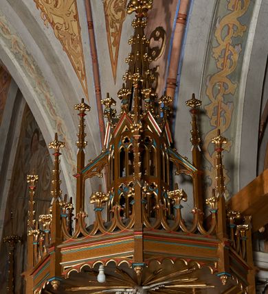Zdjęcie nr 5: Ambona przyścienna, składająca się z kosza, zaplecka i baldachimu, z dostępem od prawej strony w postaci jednobiegowych schodów z balustradą. Kosz na planie ośmioboku, ścianki dekorowane płycinami wydzielonymi przez półkolumny o kapitelach z dekoracją kwiatową na wielobocznych postumentach podtrzymujących belkowanie, którego wysuniety gzyms stanowi parapet. W polach blendy z płaskorzeźbionymi przedstawieniami czterech ewangelistów. Każdy z nich został ukazany całopostaciowo, w długiej szacie, z księgą lub zwojem w ręce i w towarzystwie symbolu apokaliptycznego. W przyłuczach blend ostrokrzew. Kosz z podwieszeniem z guzów, oparty na ośmiobocznym filarze zwieńczonym pierścieniem z dekoracją w formie rozet, ustawiony na profilowanym cokole. Balustrada dekorowana ostrołukowymi blendami. Zaplecek w kształcie wydłużonego stojącego prostokąta, półokrągły. Dekorowany płyciną z parą blend i czworoliściem z krzyżem równoramiennym. Baldachim ośmioboczny, zwieńczony ażurową wieżyczką z wimpergami, wspartą czterema przyporami, dekorowaną pinaklami, żabkami i astwerkiem; zwieńczoną pinaklem z krzyżem. Na podniebiu płaskorzeźbione przedstawienie gołębicy Ducha Świętego w glorii promienistej. Całość struktury w odcieniu ciemnego drewna, profile i detale niebieskie i czerwone, reszta profilów, wnęki blend z płaskorzeźbami i detale złocone. 