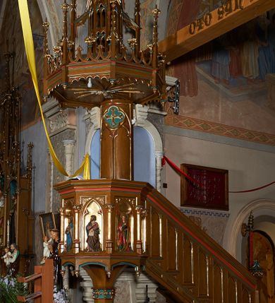 Zdjęcie nr 3: Ambona przyścienna, składająca się z kosza, zaplecka i baldachimu, z dostępem od prawej strony w postaci jednobiegowych schodów z balustradą. Kosz na planie ośmioboku, ścianki dekorowane płycinami wydzielonymi przez półkolumny o kapitelach z dekoracją kwiatową na wielobocznych postumentach podtrzymujących belkowanie, którego wysuniety gzyms stanowi parapet. W polach blendy z płaskorzeźbionymi przedstawieniami czterech ewangelistów. Każdy z nich został ukazany całopostaciowo, w długiej szacie, z księgą lub zwojem w ręce i w towarzystwie symbolu apokaliptycznego. W przyłuczach blend ostrokrzew. Kosz z podwieszeniem z guzów, oparty na ośmiobocznym filarze zwieńczonym pierścieniem z dekoracją w formie rozet, ustawiony na profilowanym cokole. Balustrada dekorowana ostrołukowymi blendami. Zaplecek w kształcie wydłużonego stojącego prostokąta, półokrągły. Dekorowany płyciną z parą blend i czworoliściem z krzyżem równoramiennym. Baldachim ośmioboczny, zwieńczony ażurową wieżyczką z wimpergami, wspartą czterema przyporami, dekorowaną pinaklami, żabkami i astwerkiem; zwieńczoną pinaklem z krzyżem. Na podniebiu płaskorzeźbione przedstawienie gołębicy Ducha Świętego w glorii promienistej. Całość struktury w odcieniu ciemnego drewna, profile i detale niebieskie i czerwone, reszta profilów, wnęki blend z płaskorzeźbami i detale złocone. 