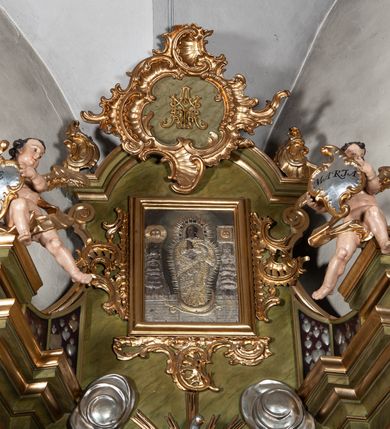 Zdjęcie nr 2: Nastawa ołtarzowa jednokondygnacyjna, jednoosiowa, ze zwieńczeniem i uszakami ustawiona na wysokim, dwudzielnym cokole. Przed nim prostopadłościenna mensa ołtarzowa z malowanym antepedium w kształcie leżącego prostokąta z przedstawieniem Ucieczki do Egiptu wzorowanym na kompozycji Giotta di Bondone. Na mensie prostopadłościenne tabernakulum zwieńczone gzymsem z drzwiczkami w kształcie stojącego prostokąta zamkniętego łukiem pełnym dekorowanymi kielichem z hostią. Kondygnacja nastawy flankowana jest przez ustawione skośnie do zewnątrz, zdwojone filary i stojące przed nimi, pozłacane kolumny o głowicach kompozytowych. W części centralnej znajduje się wnęka otoczona złotą, profilowaną ramą z rokokowym ornamentem, a w niej obraz Matki Boskiej z Dzieciątkiem, wymiennie ze sceną Zwiastowania, adorowany przez dwie flankujące go rzeźby – świętej zakonnicy po lewej i świętego zakonnika z księgą po prawej stronie. Poniżej pola obrazowego rama w kształcie leżącego prostokąta, po dwóch stronach kondygnacji uszaki w formie gablot w kształcie zbliżonym do trapezu, o wklęsło-wypukłych bokach. Wszystkie trzy wypełniają wota dziękczynne należące do obrazu Matki Boskiej. Kolumny i filary dźwigają belkowanie z rocaille'owym ornamentem na fryzie, zakończone mocno wysuniętym gzymsem, na którym umieszczono fragmenty przerwanego przyczółka. Na nim posadowiono putta trzymające kartusze, z napisami: „AVE” po lewej stronie i „MARIA” po prawej. Nad polem kondygnacji, na wysokości belkowania i gzymsu, znajduje się rzeźba gołębicy Ducha Świętego otoczona złotymi promieniami i czterema obłokami. Zwieńczenie zamknięte łukiem nadwieszonym mieści obraz z przedstawieniem Matki Boskiej Niepokalanie Poczętej w złotej profilowanej ramie, otoczony z trzech stron bujnym rocaille’owym ornamentem. Całość wieńczy kartusz utworzony z ornamentu rocaille’owego z monogramem maryjnym. Struktura polichromowana w imitacje marmoryzacji w kolorze zielonym, profile, detale i ornamenty złocone i srebrzone. 