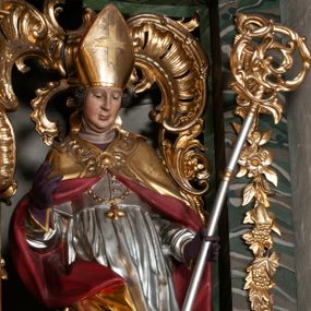 Zdjęcie nr 1: Rzeźba całopostaciowa, na cokole. Postać ukazana w lekkim skręcie, w kontrapoście, z prawą nogą wysuniętą do boku, o esowato wygiętej sylwetce. W lewej ręce trzyma pastorał, prawą unosi w geście błogosławieństwa. Głowa delikatnie opuszczona, spogląda w dół. Włosy w postaci krótkich fal zaczesane do tyłu. Twarz ma pełną, o wyraźnych rysach, małych oczach, dużym nosie i szerokich ustach. Na głowie infuła dekorowana krzyżem. Święty w stroju biskupim: sutannie, rokiecie i spiętej pod szyją kapie, pod którą ma założony pektorał, w rękawiczkach; na nogach buty. Szaty ekspresyjnie poruszone, odzwierciedlające układ sylwetki, w formie drobnych, głęboko rzeźbionych fałd. Płaszcz kontrastowo łamie się ostro wokół prawej ręki. Przy lewej nodze świętego naga półpostać Piotrowina, który wznosi ku niemu ręce. Głowę i plecy zakrywa mu częściowo biały całun. Partie ciała polichromowane naturalistycznie, sutanna, i rękawiczki fioletowe, komża biała ze złotą koronką, kapa złocona, podbita czerwienią, infuła i pektorał złote. 