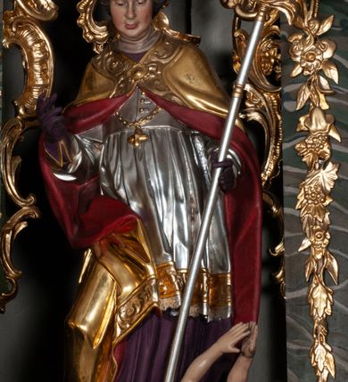 Zdjęcie nr 3: Rzeźba całopostaciowa, na cokole. Postać ukazana w lekkim skręcie, w kontrapoście, z prawą nogą wysuniętą do boku, o esowato wygiętej sylwetce. W lewej ręce trzyma pastorał, prawą unosi w geście błogosławieństwa. Głowa delikatnie opuszczona, spogląda w dół. Włosy w postaci krótkich fal zaczesane do tyłu. Twarz ma pełną, o wyraźnych rysach, małych oczach, dużym nosie i szerokich ustach. Na głowie infuła dekorowana krzyżem. Święty w stroju biskupim: sutannie, rokiecie i spiętej pod szyją kapie, pod którą ma założony pektorał, w rękawiczkach; na nogach buty. Szaty ekspresyjnie poruszone, odzwierciedlające układ sylwetki, w formie drobnych, głęboko rzeźbionych fałd. Płaszcz kontrastowo łamie się ostro wokół prawej ręki. Przy lewej nodze świętego naga półpostać Piotrowina, który wznosi ku niemu ręce. Głowę i plecy zakrywa mu częściowo biały całun. Partie ciała polichromowane naturalistycznie, sutanna, i rękawiczki fioletowe, komża biała ze złotą koronką, kapa złocona, podbita czerwienią, infuła i pektorał złote. 