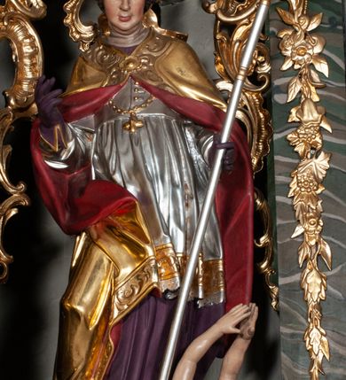 Zdjęcie nr 2: Rzeźba całopostaciowa, na cokole. Postać ukazana w lekkim skręcie, w kontrapoście, z prawą nogą wysuniętą do boku, o esowato wygiętej sylwetce. W lewej ręce trzyma pastorał, prawą unosi w geście błogosławieństwa. Głowa delikatnie opuszczona, spogląda w dół. Włosy w postaci krótkich fal zaczesane do tyłu. Twarz ma pełną, o wyraźnych rysach, małych oczach, dużym nosie i szerokich ustach. Na głowie infuła dekorowana krzyżem. Święty w stroju biskupim: sutannie, rokiecie i spiętej pod szyją kapie, pod którą ma założony pektorał, w rękawiczkach; na nogach buty. Szaty ekspresyjnie poruszone, odzwierciedlające układ sylwetki, w formie drobnych, głęboko rzeźbionych fałd. Płaszcz kontrastowo łamie się ostro wokół prawej ręki. Przy lewej nodze świętego naga półpostać Piotrowina, który wznosi ku niemu ręce. Głowę i plecy zakrywa mu częściowo biały całun. Partie ciała polichromowane naturalistycznie, sutanna, i rękawiczki fioletowe, komża biała ze złotą koronką, kapa złocona, podbita czerwienią, infuła i pektorał złote. 