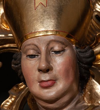 Zdjęcie nr 8: Rzeźba całopostaciowa, na cokole. Postać ukazana w lekkim skręcie, w kontrapoście, z prawą nogą wysuniętą do boku, o esowato wygiętej sylwetce. W lewej ręce trzyma pastorał, prawą unosi w geście błogosławieństwa. Głowa delikatnie opuszczona, spogląda w dół. Włosy w postaci krótkich fal zaczesane do tyłu. Twarz ma pełną, o wyraźnych rysach, małych oczach, dużym nosie i szerokich ustach. Na głowie infuła dekorowana krzyżem. Święty w stroju biskupim: sutannie, rokiecie i spiętej pod szyją kapie, pod którą ma założony pektorał, w rękawiczkach; na nogach buty. Szaty ekspresyjnie poruszone, odzwierciedlające układ sylwetki, w formie drobnych, głęboko rzeźbionych fałd. Płaszcz kontrastowo łamie się ostro wokół prawej ręki. Przy lewej nodze świętego naga półpostać Piotrowina, który wznosi ku niemu ręce. Głowę i plecy zakrywa mu częściowo biały całun. Partie ciała polichromowane naturalistycznie, sutanna, i rękawiczki fioletowe, komża biała ze złotą koronką, kapa złocona, podbita czerwienią, infuła i pektorał złote. 