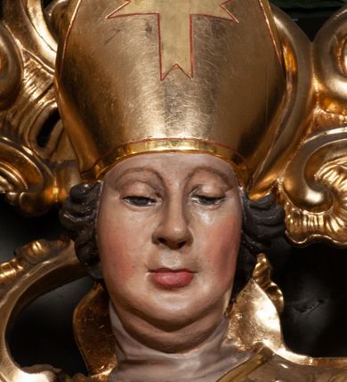 Zdjęcie nr 7: Rzeźba całopostaciowa, na cokole. Postać ukazana w lekkim skręcie, w kontrapoście, z prawą nogą wysuniętą do boku, o esowato wygiętej sylwetce. W lewej ręce trzyma pastorał, prawą unosi w geście błogosławieństwa. Głowa delikatnie opuszczona, spogląda w dół. Włosy w postaci krótkich fal zaczesane do tyłu. Twarz ma pełną, o wyraźnych rysach, małych oczach, dużym nosie i szerokich ustach. Na głowie infuła dekorowana krzyżem. Święty w stroju biskupim: sutannie, rokiecie i spiętej pod szyją kapie, pod którą ma założony pektorał, w rękawiczkach; na nogach buty. Szaty ekspresyjnie poruszone, odzwierciedlające układ sylwetki, w formie drobnych, głęboko rzeźbionych fałd. Płaszcz kontrastowo łamie się ostro wokół prawej ręki. Przy lewej nodze świętego naga półpostać Piotrowina, który wznosi ku niemu ręce. Głowę i plecy zakrywa mu częściowo biały całun. Partie ciała polichromowane naturalistycznie, sutanna, i rękawiczki fioletowe, komża biała ze złotą koronką, kapa złocona, podbita czerwienią, infuła i pektorał złote. 