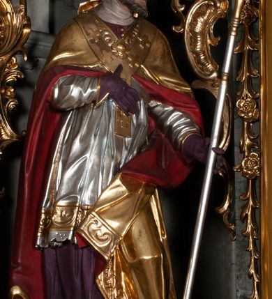 Zdjęcie nr 3: Rzeźba całopostaciowa, na cokole. Postać ukazana w skręcie, w kontrapoście, z lewą nogą odciążoną. W lewej ręce trzyma krzyż arcybiskupi, prawą rękę przyciska do piersi. Głowa zwrócona  w lewo, twarz o wyraźnych rysach, okolona krótką brodą, włosy zaczesane do tyłu. Święty ma brązowe oczy, wydatny nos i lekko rozchylone usta. Przedstawiono go w stroju biskupim: sutannie, rokiecie i spiętej pod szyją kapie, na którą nałożono paliusz i w rękawiczkach; na nogach buty. Szaty odzwierciedlają układ sylwetki układając się w drobne, głęboko rzeźbione fałdy. Płaszcz kontrastowo łamie się ostro wokół lewej ręki; spod szat widać czubki butów. Partie ciała polichromowane naturalistycznie, sutanna, i rękawiczki fioletowe, komża biała ze złotą koronką, kapa złocona, podbita czerwienią, infuła i paliusz złote.