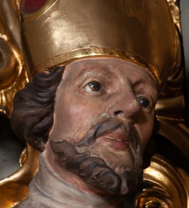 Zdjęcie nr 6: Rzeźba całopostaciowa, na cokole. Postać ukazana w skręcie, w kontrapoście, z lewą nogą odciążoną. W lewej ręce trzyma krzyż arcybiskupi, prawą rękę przyciska do piersi. Głowa zwrócona  w lewo, twarz o wyraźnych rysach, okolona krótką brodą, włosy zaczesane do tyłu. Święty ma brązowe oczy, wydatny nos i lekko rozchylone usta. Przedstawiono go w stroju biskupim: sutannie, rokiecie i spiętej pod szyją kapie, na którą nałożono paliusz i w rękawiczkach; na nogach buty. Szaty odzwierciedlają układ sylwetki układając się w drobne, głęboko rzeźbione fałdy. Płaszcz kontrastowo łamie się ostro wokół lewej ręki; spod szat widać czubki butów. Partie ciała polichromowane naturalistycznie, sutanna, i rękawiczki fioletowe, komża biała ze złotą koronką, kapa złocona, podbita czerwienią, infuła i paliusz złote.