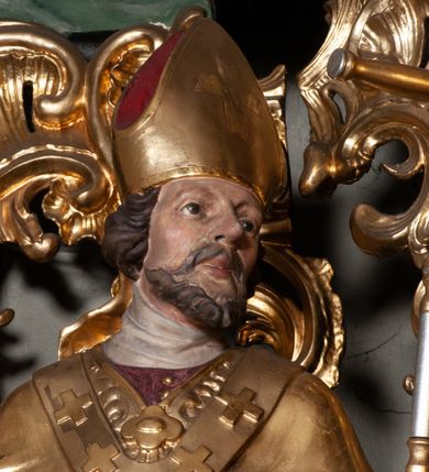 Zdjęcie nr 5: Rzeźba całopostaciowa, na cokole. Postać ukazana w skręcie, w kontrapoście, z lewą nogą odciążoną. W lewej ręce trzyma krzyż arcybiskupi, prawą rękę przyciska do piersi. Głowa zwrócona  w lewo, twarz o wyraźnych rysach, okolona krótką brodą, włosy zaczesane do tyłu. Święty ma brązowe oczy, wydatny nos i lekko rozchylone usta. Przedstawiono go w stroju biskupim: sutannie, rokiecie i spiętej pod szyją kapie, na którą nałożono paliusz i w rękawiczkach; na nogach buty. Szaty odzwierciedlają układ sylwetki układając się w drobne, głęboko rzeźbione fałdy. Płaszcz kontrastowo łamie się ostro wokół lewej ręki; spod szat widać czubki butów. Partie ciała polichromowane naturalistycznie, sutanna, i rękawiczki fioletowe, komża biała ze złotą koronką, kapa złocona, podbita czerwienią, infuła i paliusz złote.