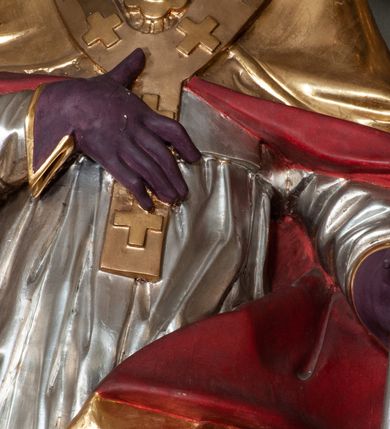 Zdjęcie nr 7: Rzeźba całopostaciowa, na cokole. Postać ukazana w skręcie, w kontrapoście, z lewą nogą odciążoną. W lewej ręce trzyma krzyż arcybiskupi, prawą rękę przyciska do piersi. Głowa zwrócona  w lewo, twarz o wyraźnych rysach, okolona krótką brodą, włosy zaczesane do tyłu. Święty ma brązowe oczy, wydatny nos i lekko rozchylone usta. Przedstawiono go w stroju biskupim: sutannie, rokiecie i spiętej pod szyją kapie, na którą nałożono paliusz i w rękawiczkach; na nogach buty. Szaty odzwierciedlają układ sylwetki układając się w drobne, głęboko rzeźbione fałdy. Płaszcz kontrastowo łamie się ostro wokół lewej ręki; spod szat widać czubki butów. Partie ciała polichromowane naturalistycznie, sutanna, i rękawiczki fioletowe, komża biała ze złotą koronką, kapa złocona, podbita czerwienią, infuła i paliusz złote.