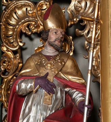 Zdjęcie nr 1: Rzeźba całopostaciowa, na cokole. Postać ukazana w skręcie, w kontrapoście, z lewą nogą odciążoną. W lewej ręce trzyma krzyż arcybiskupi, prawą rękę przyciska do piersi. Głowa zwrócona  w lewo, twarz o wyraźnych rysach, okolona krótką brodą, włosy zaczesane do tyłu. Święty ma brązowe oczy, wydatny nos i lekko rozchylone usta. Przedstawiono go w stroju biskupim: sutannie, rokiecie i spiętej pod szyją kapie, na którą nałożono paliusz i w rękawiczkach; na nogach buty. Szaty odzwierciedlają układ sylwetki układając się w drobne, głęboko rzeźbione fałdy. Płaszcz kontrastowo łamie się ostro wokół lewej ręki; spod szat widać czubki butów. Partie ciała polichromowane naturalistycznie, sutanna, i rękawiczki fioletowe, komża biała ze złotą koronką, kapa złocona, podbita czerwienią, infuła i paliusz złote.