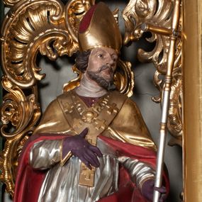 Zdjęcie nr 1: Rzeźba całopostaciowa, na cokole. Postać ukazana w skręcie, w kontrapoście, z lewą nogą odciążoną. W lewej ręce trzyma krzyż arcybiskupi, prawą rękę przyciska do piersi. Głowa zwrócona  w lewo, twarz o wyraźnych rysach, okolona krótką brodą, włosy zaczesane do tyłu. Święty ma brązowe oczy, wydatny nos i lekko rozchylone usta. Przedstawiono go w stroju biskupim: sutannie, rokiecie i spiętej pod szyją kapie, na którą nałożono paliusz i w rękawiczkach; na nogach buty. Szaty odzwierciedlają układ sylwetki układając się w drobne, głęboko rzeźbione fałdy. Płaszcz kontrastowo łamie się ostro wokół lewej ręki; spod szat widać czubki butów. Partie ciała polichromowane naturalistycznie, sutanna, i rękawiczki fioletowe, komża biała ze złotą koronką, kapa złocona, podbita czerwienią, infuła i paliusz złote.