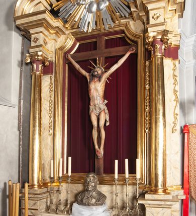 Zdjęcie nr 1: Ołtarz jednokondygnacyjny, jednoosiowy, na wysokim cokole, ze zwieńczeniem w formie glorii. Mensa ołtarzowa w kształcie prostopadłościanu, antepedium w postaci leżącego prostokąta, tkane, dekorowane rzędem maków w dolnej partii. Kondygnacja nastawy flankowana przez zwielokrotnione pilastry, ozdobione z zewnętrznych stron ornamentami roślinnymi i wysuniętymi przed nie kolumnami z głowicami kompozytowymi, ozdobionymi wolutami i liśćmi akantu. Kolumny i pilastry ustawione na wysokim cokole dekorowanym płycinami, dźwigają rozbudowane belkowanie, w pasie fryzu dekorowane rozetami. Belkowanie przełamuje się nad zwielokrotnionymi pilastrami i kolumnami i podtrzymuje fragment przerwanego przyczółka, na którym siedzą dwa putta. W polu znajduje się wnęka w kształcie stojącego prostokąta o górnych narożach wklęsłych, w której umieszczono krucyfiks. Zwieńczenie tworzy gloria promienista z centralnie umieszczonym okiem Opatrzności w otoku z obłoków i główek anielskich. Struktura polichromowana, marmoryzowana w odcieniu kremowym, kolumny, ornamenty i profile złocone, płyciny bordowe, gloria srebrzona i złocona.  