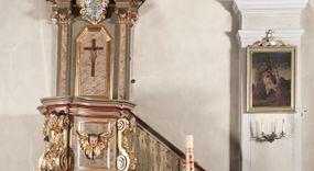 Zdjęcie nr 1: Ambona przyścienna, wisząca, dostępna od strony prezbiterium. Kosz na planie wklęsło-wypukłym, na  zwężającym się cokole dekorowanym liśćmi. U spodu podwieszone dekorowane rocaille’m woluty podtrzymujące szyszkę. Ścianki kosza wklęsłe, zdobione  płycinami wypełnionymi główkami anielskimi, a ścięte naroża wolutami z rocaille’m i główkami anielskimi. Kosz zamknięty parapetem w formie belkowania, wyłamanego na narożach. Z prawej strony do kosza prowadzą schody z ażurową balustradą wyciętą w pionowe, fantazyjne kształty, z poręczą. Zaplecek w kształcie stojącego prostokąta flankowany pilastrami wypełnionymi płycinami z głowicami utworzonymi z wolut i główek anielskich, podtrzymującymi fragmenty pełnego, wyłamanego na ich osiach belkowania. W polu płycina w kształcie stojącego prostokąta o ściętych górnych narożach z krucyfiksem. Nad nią, między fragmentami belkowania pięciopolowy herb Wazów zwieńczony koroną. Ponad zapleckiem płaskorzeźbiona gołębica Ducha Świętego w otoku z obłoków na tle dużych rozmiarów glorii promienistej. 
Struktura polichromowana w kolorze zielonym; płyciny marmoryzowane w odcieniu jasnego brązu; profilowania brązowe, detale, ornamenty, gloria złocone, gołębica i obłoki srebrzone. 

