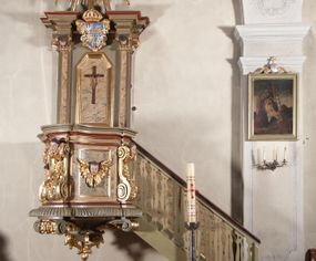 Zdjęcie nr 1: Ambona przyścienna, wisząca, dostępna od strony prezbiterium. Kosz na planie wklęsło-wypukłym, na  zwężającym się cokole dekorowanym liśćmi. U spodu podwieszone dekorowane rocaille’m woluty podtrzymujące szyszkę. Ścianki kosza wklęsłe, zdobione  płycinami wypełnionymi główkami anielskimi, a ścięte naroża wolutami z rocaille’m i główkami anielskimi. Kosz zamknięty parapetem w formie belkowania, wyłamanego na narożach. Z prawej strony do kosza prowadzą schody z ażurową balustradą wyciętą w pionowe, fantazyjne kształty, z poręczą. Zaplecek w kształcie stojącego prostokąta flankowany pilastrami wypełnionymi płycinami z głowicami utworzonymi z wolut i główek anielskich, podtrzymującymi fragmenty pełnego, wyłamanego na ich osiach belkowania. W polu płycina w kształcie stojącego prostokąta o ściętych górnych narożach z krucyfiksem. Nad nią, między fragmentami belkowania pięciopolowy herb Wazów zwieńczony koroną. Ponad zapleckiem płaskorzeźbiona gołębica Ducha Świętego w otoku z obłoków na tle dużych rozmiarów glorii promienistej. 
Struktura polichromowana w kolorze zielonym; płyciny marmoryzowane w odcieniu jasnego brązu; profilowania brązowe, detale, ornamenty, gloria złocone, gołębica i obłoki srebrzone. 
