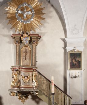 Zdjęcie nr 1: Ambona przyścienna, wisząca, dostępna od strony prezbiterium. Kosz na planie wklęsło-wypukłym, na  zwężającym się cokole dekorowanym liśćmi. U spodu podwieszone dekorowane rocaille’m woluty podtrzymujące szyszkę. Ścianki kosza wklęsłe, zdobione  płycinami wypełnionymi główkami anielskimi, a ścięte naroża wolutami z rocaille’m i główkami anielskimi. Kosz zamknięty parapetem w formie belkowania, wyłamanego na narożach. Z prawej strony do kosza prowadzą schody z ażurową balustradą wyciętą w pionowe, fantazyjne kształty, z poręczą. Zaplecek w kształcie stojącego prostokąta flankowany pilastrami wypełnionymi płycinami z głowicami utworzonymi z wolut i główek anielskich, podtrzymującymi fragmenty pełnego, wyłamanego na ich osiach belkowania. W polu płycina w kształcie stojącego prostokąta o ściętych górnych narożach z krucyfiksem. Nad nią, między fragmentami belkowania pięciopolowy herb Wazów zwieńczony koroną. Ponad zapleckiem płaskorzeźbiona gołębica Ducha Świętego w otoku z obłoków na tle dużych rozmiarów glorii promienistej. 
Struktura polichromowana w kolorze zielonym; płyciny marmoryzowane w odcieniu jasnego brązu; profilowania brązowe, detale, ornamenty, gloria złocone, gołębica i obłoki srebrzone. 
