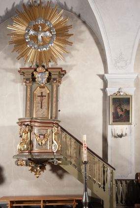 Zdjęcie nr 1: Ambona przyścienna, wisząca, dostępna od strony prezbiterium. Kosz na planie wklęsło-wypukłym, na  zwężającym się cokole dekorowanym liśćmi. U spodu podwieszone dekorowane rocaille’m woluty podtrzymujące szyszkę. Ścianki kosza wklęsłe, zdobione  płycinami wypełnionymi główkami anielskimi, a ścięte naroża wolutami z rocaille’m i główkami anielskimi. Kosz zamknięty parapetem w formie belkowania, wyłamanego na narożach. Z prawej strony do kosza prowadzą schody z ażurową balustradą wyciętą w pionowe, fantazyjne kształty, z poręczą. Zaplecek w kształcie stojącego prostokąta flankowany pilastrami wypełnionymi płycinami z głowicami utworzonymi z wolut i główek anielskich, podtrzymującymi fragmenty pełnego, wyłamanego na ich osiach belkowania. W polu płycina w kształcie stojącego prostokąta o ściętych górnych narożach z krucyfiksem. Nad nią, między fragmentami belkowania pięciopolowy herb Wazów zwieńczony koroną. Ponad zapleckiem płaskorzeźbiona gołębica Ducha Świętego w otoku z obłoków na tle dużych rozmiarów glorii promienistej. 
Struktura polichromowana w kolorze zielonym; płyciny marmoryzowane w odcieniu jasnego brązu; profilowania brązowe, detale, ornamenty, gloria złocone, gołębica i obłoki srebrzone. 
