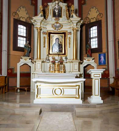Zdjęcie nr 19: Ołtarz architektoniczny, drewniany, jednoosiowy, jednokondygnacyjny z bramkami. Mensa wąska, prostopadłościenna o ściankach wklęsło-wypukłych. Tabernakulum w kształcie prostopadłościennej skrzynki z przodu o linii wklęsło-wypukłej z uskokami. Ustawione jest na  wysokim cokole, flankowane parą szerokich, wklęsłych pilastrów, zdobionych ornamentem złożonym z przeplatających się wolut i kampanul. Ścianka frontowa wypukła, w polu prostokątne, metalowe drzwiczki, zdobione kiśćmi winogron i kłosami zbóż, pośrodku łaciński prosty krzyż. Całość zamknięta profilowanym gzymsem. Po bokach tabernakulum zwisy z kampanul, a na skrajach płaskorzeźbione uszy z liści akantu.  Do górnej krawędzi przybite gwoździem na trzech tulejach lampki oliwne. Nad tabernakulum tron wystawienia z rzeźbionymi w drewnie aniołkami na obłokach adorującymi krucyfiks. 

Retabulum ustawione na zdwojonym cokole, ujęte zwielokrotnionymi pilastrami toskańskimi i dwiema kolumnami kompozytowymi na wysokich cokołach, ustawionymi pod kątem i podtrzymującymi profilowane, przerwane belkowanie. Pilastry od strony wewnętrznej ołtarza są wklęsłe, ozdobione dwiema parami uskrzydlonych główek anielskich i zwisami z kwiatów; od zewnątrz pilastry mają trzony wypukłe.  W polu głównym wnęka, ujęta ramą w kształcie stojącego prostokąta o uszatych narożach. We wnęce na czerwonym tle umieszczony obraz Matki Boskiej Piekarskiej, na zasuwach obrazy „Koronacja Matki Boskiej” oraz „Św. Mikołaj”. Nad obrazem kartusz o kształcie zbliżonym do rombu z ornamentami rocaille&#039;owymi wokół; w polu napis „ALTARE / PRIVILEGI/ATUM”. Na osi kolumn, na przyczółku ustawione są wazony z kwiatami. 

Zwieńczenie w kształcie stojącego prostokąta, flankowane parą pilastrów podtrzymujących przerwane belkowanie, zamknięte gzymsem w formie łuku odcinkowego. Na szczycie promienista gloria z otokiem chmur pośrodku z płaskorzeźbionym monogramem IHS. W polu głównym obraz przedstawiający Ekstazę św. Teresy w kształcie stojącego prostokąta, zamknięty łukiem nadwieszonym. Po bokach zwieńczenia uszy z liści akantu i kwiatów. Pod obrazem uskrzydlona para główek aniołków. 

Bramki w formie stojącego prostokąta, flankowane węgarami, zamknięte wolutowym łukiem z ustawionymi na szczycie na niskich cokołach rzeźbami świętych: po lewej św. Piotra, a po prawej św. Pawła.  Przejście zamknięte łukiem wklęsło-wypukłym z uskokami. Struktura polichromowana w kolorze jasnobeżowym, ornamentyka złocona, detale srebrzone.

