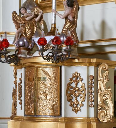 Zdjęcie nr 14: Ołtarz architektoniczny, drewniany, jednoosiowy, jednokondygnacyjny z bramkami. Mensa wąska, prostopadłościenna o ściankach wklęsło-wypukłych. Tabernakulum w kształcie prostopadłościennej skrzynki z przodu o linii wklęsło-wypukłej z uskokami. Ustawione jest na  wysokim cokole, flankowane parą szerokich, wklęsłych pilastrów, zdobionych ornamentem złożonym z przeplatających się wolut i kampanul. Ścianka frontowa wypukła, w polu prostokątne, metalowe drzwiczki, zdobione kiśćmi winogron i kłosami zbóż, pośrodku łaciński prosty krzyż. Całość zamknięta profilowanym gzymsem. Po bokach tabernakulum zwisy z kampanul, a na skrajach płaskorzeźbione uszy z liści akantu.  Do górnej krawędzi przybite gwoździem na trzech tulejach lampki oliwne. Nad tabernakulum tron wystawienia z rzeźbionymi w drewnie aniołkami na obłokach adorującymi krucyfiks. 

Retabulum ustawione na zdwojonym cokole, ujęte zwielokrotnionymi pilastrami toskańskimi i dwiema kolumnami kompozytowymi na wysokich cokołach, ustawionymi pod kątem i podtrzymującymi profilowane, przerwane belkowanie. Pilastry od strony wewnętrznej ołtarza są wklęsłe, ozdobione dwiema parami uskrzydlonych główek anielskich i zwisami z kwiatów; od zewnątrz pilastry mają trzony wypukłe.  W polu głównym wnęka, ujęta ramą w kształcie stojącego prostokąta o uszatych narożach. We wnęce na czerwonym tle umieszczony obraz Matki Boskiej Piekarskiej, na zasuwach obrazy „Koronacja Matki Boskiej” oraz „Św. Mikołaj”. Nad obrazem kartusz o kształcie zbliżonym do rombu z ornamentami rocaille&#039;owymi wokół; w polu napis „ALTARE / PRIVILEGI/ATUM”. Na osi kolumn, na przyczółku ustawione są wazony z kwiatami. 

Zwieńczenie w kształcie stojącego prostokąta, flankowane parą pilastrów podtrzymujących przerwane belkowanie, zamknięte gzymsem w formie łuku odcinkowego. Na szczycie promienista gloria z otokiem chmur pośrodku z płaskorzeźbionym monogramem IHS. W polu głównym obraz przedstawiający Ekstazę św. Teresy w kształcie stojącego prostokąta, zamknięty łukiem nadwieszonym. Po bokach zwieńczenia uszy z liści akantu i kwiatów. Pod obrazem uskrzydlona para główek aniołków. 

Bramki w formie stojącego prostokąta, flankowane węgarami, zamknięte wolutowym łukiem z ustawionymi na szczycie na niskich cokołach rzeźbami świętych: po lewej św. Piotra, a po prawej św. Pawła.  Przejście zamknięte łukiem wklęsło-wypukłym z uskokami. Struktura polichromowana w kolorze jasnobeżowym, ornamentyka złocona, detale srebrzone.
