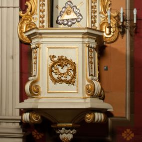 Zdjęcie nr 1: Ambona drewniana przy północnej ścianie prezbiterium; z koszem na rzucie czworoboku ze ściętymi narożami. Część dolna w formie odwróconego, ściętego ostrosłupa z podwieszonym owocem winnego grona, w kształcie kulistym, otoczonym liśćmi akantu. Na podwieszeniu, w narożach dużych rozmiarów woluty. Kosz wydzielony profilowanym cokołem u dołu. W narożach pilastry o trzonach wypełnionych podwieszonymi kampanulami; w dolnej części przechodzą w woluty. Kapitele zredukowane do małej wolutki. W polach ścian bocznych złocone, prostokątne płyciny wypełnione dekoracją rocaille'ową oraz akantem. Na licu większy kartusz utworzony z akantu i rocaille'a. Kosz wieńczy wydatny, silnie profilowany gzyms. Zaplecek prostokątny, ujęty uszakami z akantu, zakończony wolutami. Flankowany pilastrami pozbawionymi kapiteli, trzony wypełnione podwieszonymi kampanulami. Drzwi prostokątne zamknięte łukiem kotarowym ściętym, w centrum złocone oko Opatrzności na tle srebrzonych obłoków i złotej glorii promienistej. Nad drzwiami, na osi, złocona muszla palmetowa. Baldachim na rzucie analogicznym jak kosz, w podniebieniu srebrzona gołębica Ducha Świętego na tle glorii promienistej. Wokół podwieszony lambrekin o srebrzonych chwostach. Brzegi profilowane, na osi środkowej złocony kartusz rocaille'owy. Zwieńczenie w formie ściętego ostrosłupa z wolutami na narożach. Na szczycie pełnoplastyczna rzeźba anioła dmącego w trąbę, który w lewej dłoni trzyma z księgą z tekstem „RZEKŁ / BÓG / DO PANA / MEGO / SIĄDŹ / PO PRAWICY / MOJEJ”. Ambona utrzymana w kolorystyce jasnego beżu, złocenia w partiach ornamentu i załamaniach profili detalu architektonicznego.