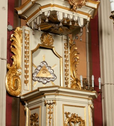 Zdjęcie nr 3: Ambona drewniana przy północnej ścianie prezbiterium; z koszem na rzucie czworoboku ze ściętymi narożami. Część dolna w formie odwróconego, ściętego ostrosłupa z podwieszonym owocem winnego grona, w kształcie kulistym, otoczonym liśćmi akantu. Na podwieszeniu, w narożach dużych rozmiarów woluty. Kosz wydzielony profilowanym cokołem u dołu. W narożach pilastry o trzonach wypełnionych podwieszonymi kampanulami; w dolnej części przechodzą w woluty. Kapitele zredukowane do małej wolutki. W polach ścian bocznych złocone, prostokątne płyciny wypełnione dekoracją rocaille'ową oraz akantem. Na licu większy kartusz utworzony z akantu i rocaille'a. Kosz wieńczy wydatny, silnie profilowany gzyms. Zaplecek prostokątny, ujęty uszakami z akantu, zakończony wolutami. Flankowany pilastrami pozbawionymi kapiteli, trzony wypełnione podwieszonymi kampanulami. Drzwi prostokątne zamknięte łukiem kotarowym ściętym, w centrum złocone oko Opatrzności na tle srebrzonych obłoków i złotej glorii promienistej. Nad drzwiami, na osi, złocona muszla palmetowa. Baldachim na rzucie analogicznym jak kosz, w podniebieniu srebrzona gołębica Ducha Świętego na tle glorii promienistej. Wokół podwieszony lambrekin o srebrzonych chwostach. Brzegi profilowane, na osi środkowej złocony kartusz rocaille'owy. Zwieńczenie w formie ściętego ostrosłupa z wolutami na narożach. Na szczycie pełnoplastyczna rzeźba anioła dmącego w trąbę, który w lewej dłoni trzyma z księgą z tekstem „RZEKŁ / BÓG / DO PANA / MEGO / SIĄDŹ / PO PRAWICY / MOJEJ”. Ambona utrzymana w kolorystyce jasnego beżu, złocenia w partiach ornamentu i załamaniach profili detalu architektonicznego.