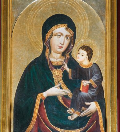 Zdjęcie nr 2: Obraz w kształcie stojącego prostokąta zamkniętego półkoliście. Matka Boska z Dzieciątkiem na ręku przedstawiona frontalnie w półpostaci, z głową lekko przechyloną na lewe ramię. Ręce ugięte w łokciach, lewą podtrzymuje Dzieciątko, wskazując na nie prawą dłonią. Twarz pełna, modelowana światłocieniowo, o dużych oczach, małych ustach i wydatnym podbródku. Cała postać Marii wraz z głową okryta maforionem ozdobionym na brzegach lamówką i spiętym na piersiach ozdobną klamrą z literami: ihs. Lamówka pierwotnie srebrzona i laserowana, ozdobiona malowaną wicią z motywów półpalmet.  Spod maforionu widoczne jedynie niewielkie fragmenty sukni ozdobione lamówką, przy dekolcie z minuskułowym napisem: maria, oraz chusta na głowie nieosłaniająca szyi. Dzieciątko siedzi na lewej ręce Marii, zwrócone w trzech czwartych w kierunku widza. Ręce ugięte w łokciach; prawą z palcami ułożonymi w geście błogosławieństwa kieruje w stronę Matki, lewą podtrzymuje kodeks, który opiera na lewym kolanie. Jezus ubrany jest w długą tunikę z długimi, szerokimi rękawami, ozdobioną przy brzegach ozdobną lamówką. Wokół głów koliste nimby z koncentrycznych okręgów. Tło złote.  