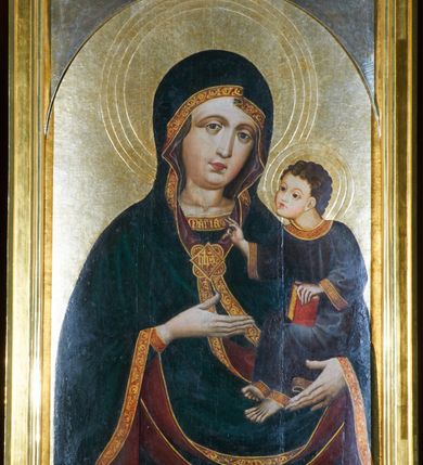 Zdjęcie nr 19: Obraz w kształcie stojącego prostokąta zamkniętego półkoliście. Matka Boska z Dzieciątkiem na ręku przedstawiona frontalnie w półpostaci, z głową lekko przechyloną na lewe ramię. Ręce ugięte w łokciach, lewą podtrzymuje Dzieciątko, wskazując na nie prawą dłonią. Twarz pełna, modelowana światłocieniowo, o dużych oczach, małych ustach i wydatnym podbródku. Cała postać Marii wraz z głową okryta maforionem ozdobionym na brzegach lamówką i spiętym na piersiach ozdobną klamrą z literami: ihs. Lamówka pierwotnie srebrzona i laserowana, ozdobiona malowaną wicią z motywów półpalmet.  Spod maforionu widoczne jedynie niewielkie fragmenty sukni ozdobione lamówką, przy dekolcie z minuskułowym napisem: maria, oraz chusta na głowie nieosłaniająca szyi. Dzieciątko siedzi na lewej ręce Marii, zwrócone w trzech czwartych w kierunku widza. Ręce ugięte w łokciach; prawą z palcami ułożonymi w geście błogosławieństwa kieruje w stronę Matki, lewą podtrzymuje kodeks, który opiera na lewym kolanie. Jezus ubrany jest w długą tunikę z długimi, szerokimi rękawami, ozdobioną przy brzegach ozdobną lamówką. Wokół głów koliste nimby z koncentrycznych okręgów. Tło złote.  