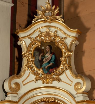 Zdjęcie nr 3: Obraz w kształcie zbliżonym do owalu, ujęty kartuszową, rocaille'ową ramą o nieregularnym, wewnętrznym obrysie ograniczającym pole obrazu. W centrum kompozycji ukazana św. Barbara w trzech czwartych, zwrócona w prawo, w prawej ręce trzyma kielich z hostią, a w lewej  miecz. Twarz owalna, z drobnymi ustami, wzrok skierowany ku górze, pucułowate policzki. Ubrana jest w bogate szaty: błękitną suknię z rękawami zakończonymi falbaną, z dużym dekoltem oraz w różowoczerwony płaszcz. Włosy długie, upięte do tyłu, z puklami opadającymi na ramiona. Po lewej stronie obrazu wieża z trzema oknami. Tło jednolite, brunatne. Kolorystka pierwszego planu żywa, tło stonowane.