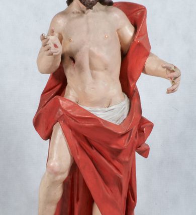 Zdjęcie nr 6: Rzeźba Chrystusa Zmartwychwstałego pełnoplastyczna, wolnostojąca, ustawiona na niskim cokole. Chrystus ukazany w całej postaci, ustawiony frontalnie, w delikatnym kontrapoście, ma prawą nogę ugiętą w kolanie i wspartą na czaszce z wężem. Ręce ma ugięte w łokciach i wyciągnięte przed siebie, w lewej trzyma chorągiew o długim drzewcu, a prawą unosi w geście błogosławieństwa. Jego twarz jest pociągła, o wysokim czole, długim i wąskim nosie, dużych oczach i rozchylonych ustach, okolona średniej długości, ciemną brodą i długimi, falowanymi włosami, które opadają na plecy i lewe ramię. Wokół głowy ma złoty nimb krzyżowy utworzony z wiązek promieni o nierównej długości. Chrystus ubrany jest w białe perizonium oraz czerwony płaszcz odsłaniający tors, otaczający figurę i przerzucony przez lewe ramię. Na dłoniach, stopach i boku zaznaczone są ślady męki. Polichromia ciała jest naturalistyczna, nimb pozłacany, drzewce chorągwi srebrzone.

